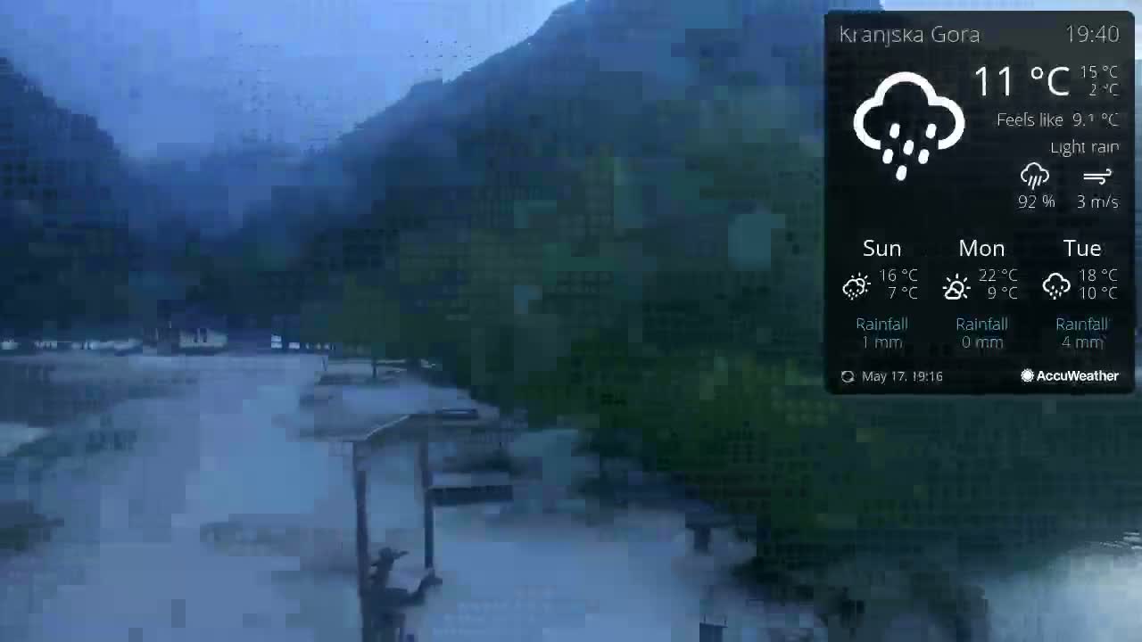 A rain-soaked, dimly lit outdoor scene shows a flat, paved area near a mountainous landscape under light rain.