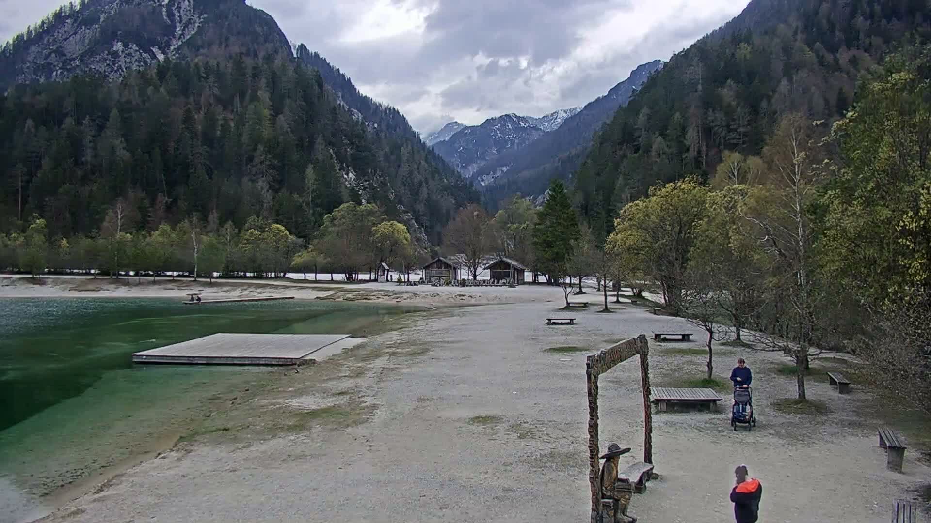 A sunny day shows a calm lake with people relaxing by the shore, boats on the water, and mountains in the background.