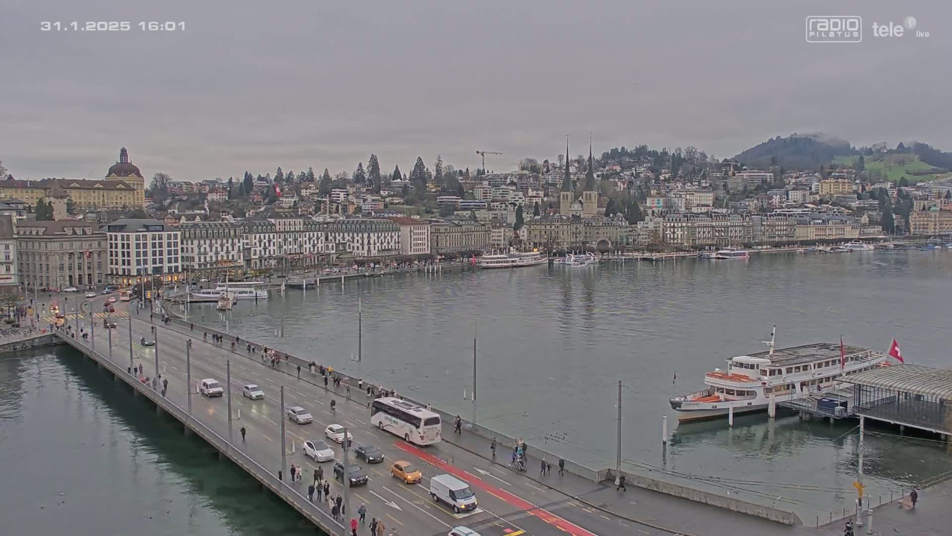 A bridge with cars and pedestrians spans a body of water, overlooking a city with overcast skies.