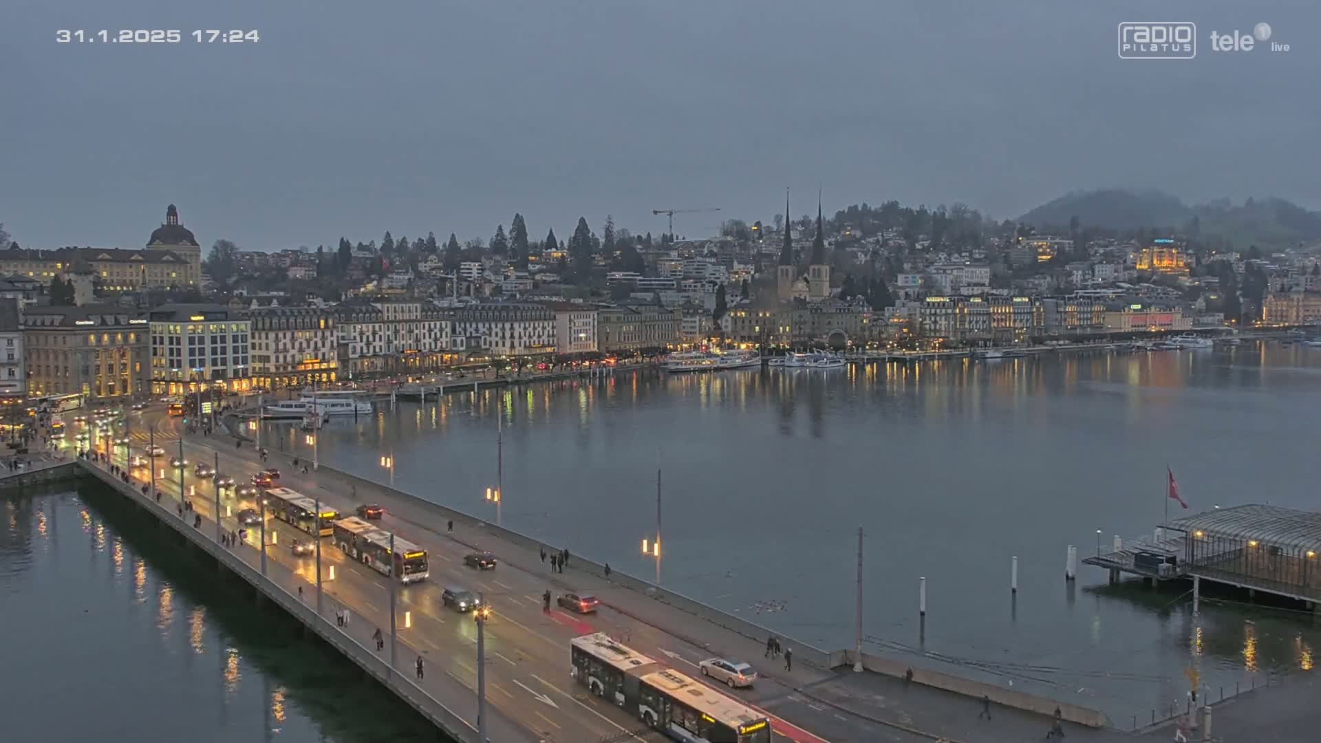 A city waterfront at dusk, with a bridge carrying traffic and buses over calm water reflecting the lights of buildings lining the shore.