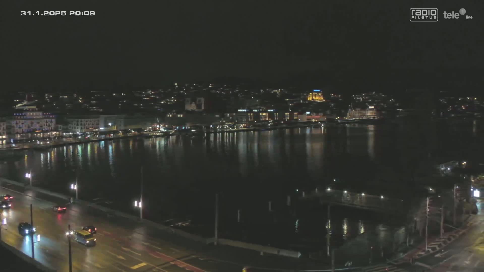 A nighttime view of a city waterfront with calm water reflecting city lights, and a road with cars driving along it.