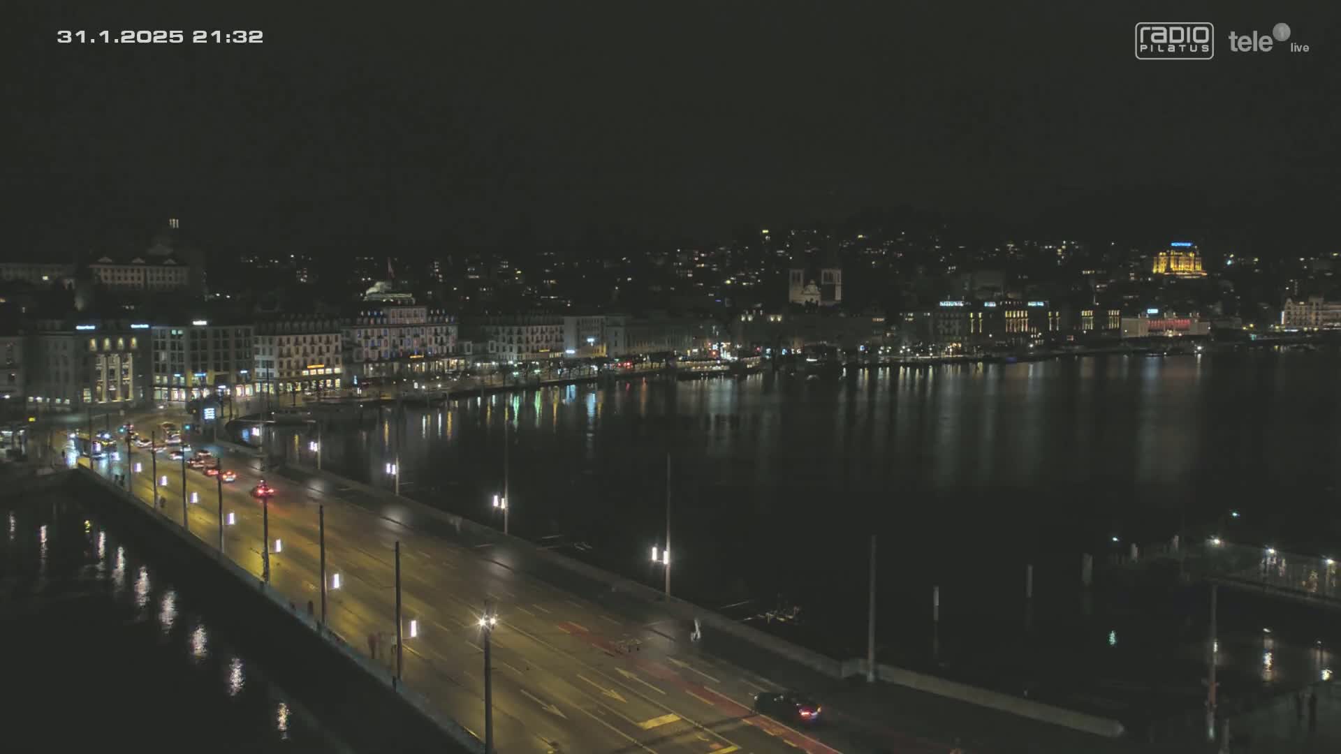 A nighttime view shows a city's waterfront with buildings lit up along the shore and a bridge with cars driving across it, all reflecting in dark water under a clear sky.