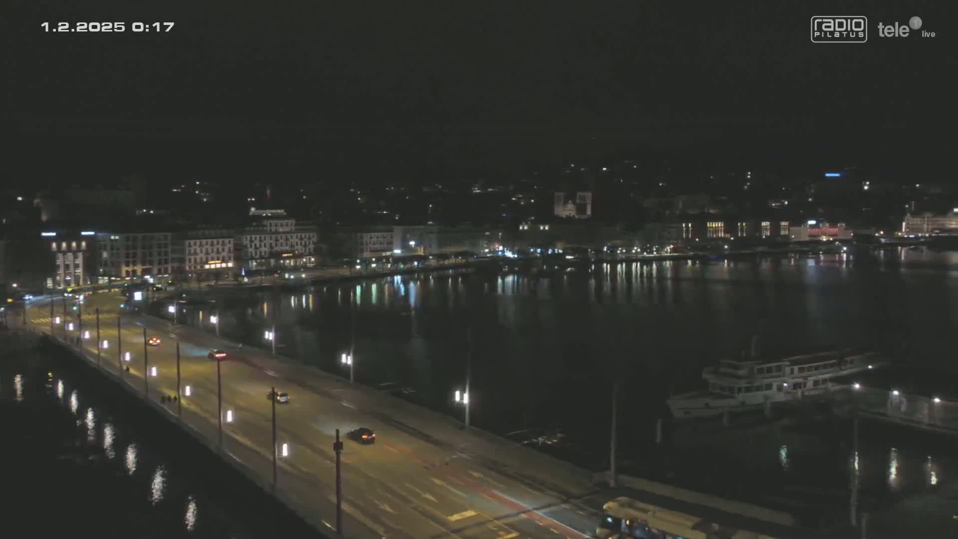 A nighttime view of a city waterfront, showing a bridge with cars driving across it, buildings lining the shore, and a boat docked in the dark water.