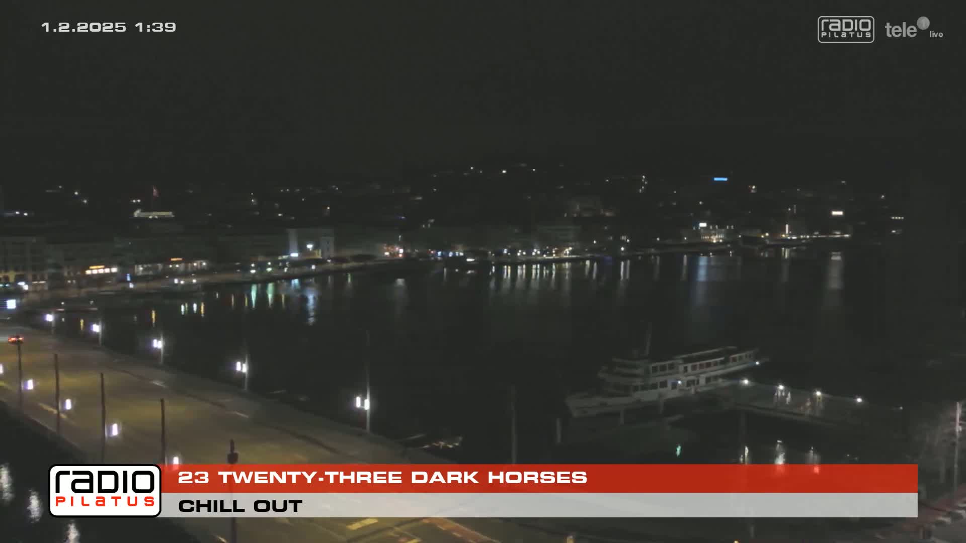 A nighttime view of a calm body of water with city lights reflecting on its surface, a boat docked near a bridge, and a dark city skyline in the background.