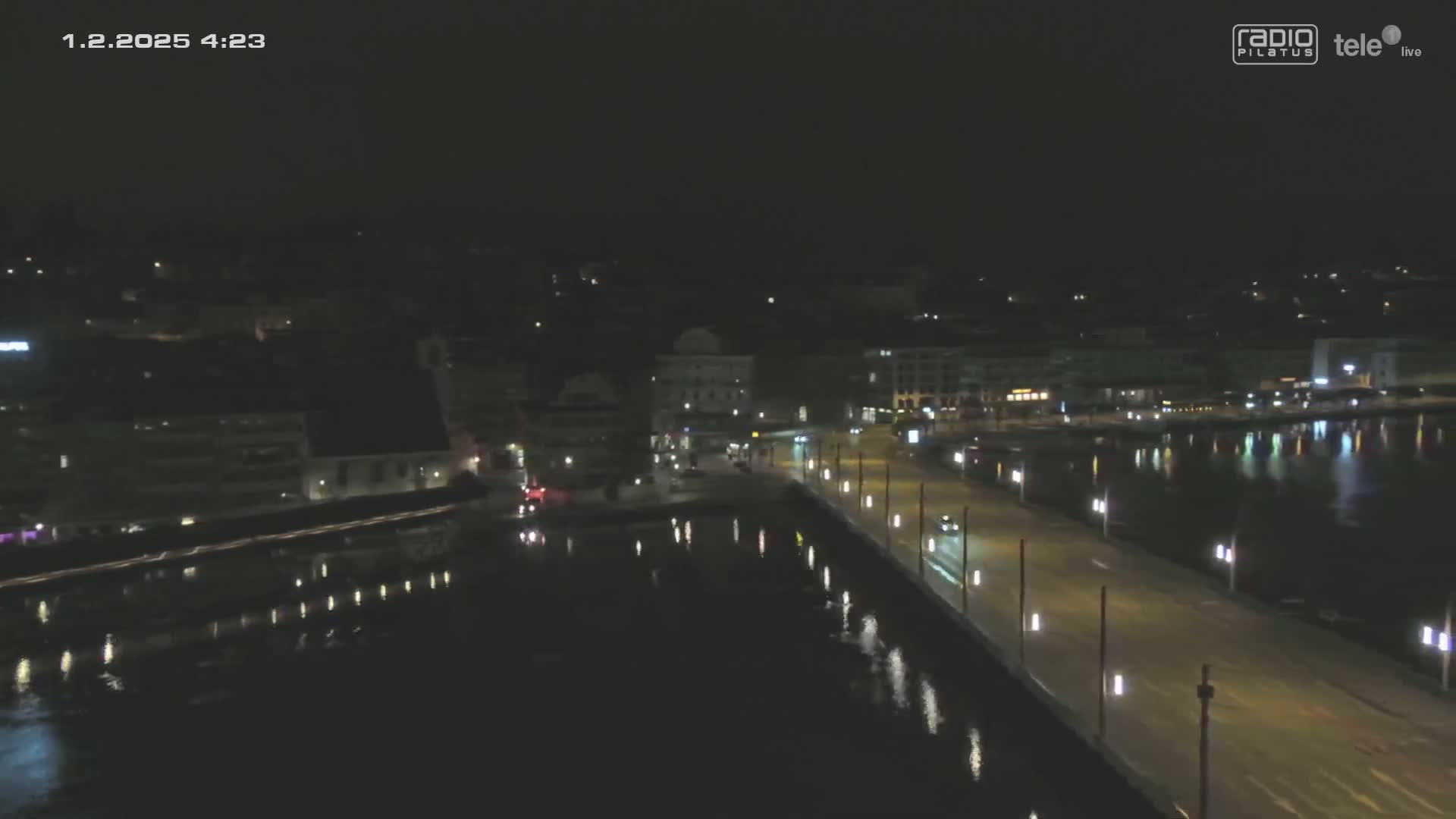 A nighttime, high-angle view shows a city waterfront with a bridge, buildings, and calm water reflecting lights.