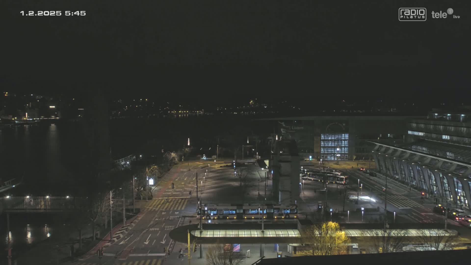 A nighttime, high-angle view of a city waterfront with a street, several buses and a tram, and a large modern building near a body of water.
