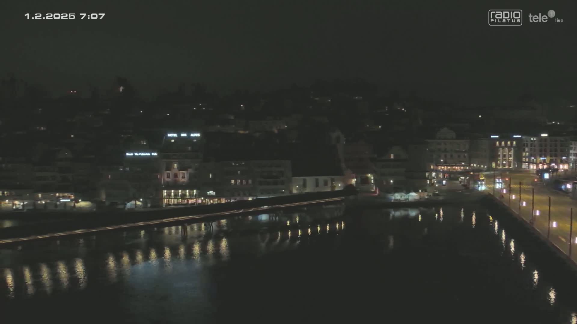 A nighttime, high-angle view of a city waterfront with a bridge and calm water reflecting the lights of buildings along the shore.