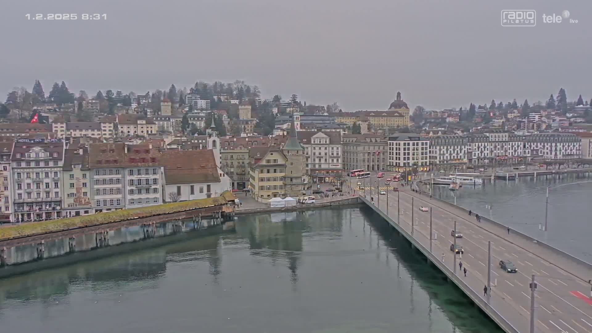 A long bridge spans a calm body of water, leading to a city of European-style buildings under an overcast sky.