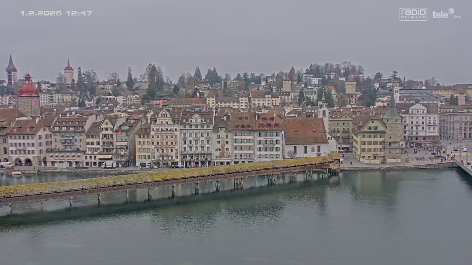 A waterfront city with numerous buildings, a long bridge, and calm water under an overcast sky.