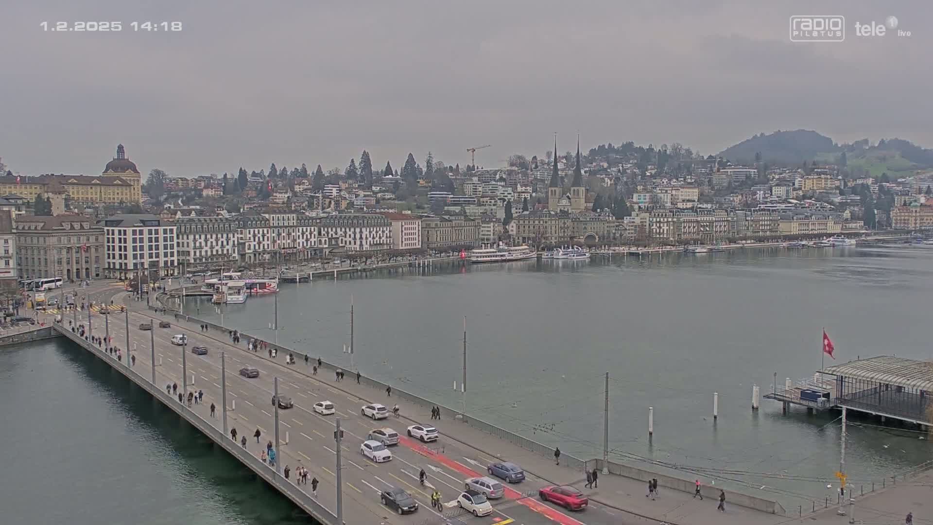 A long bridge with pedestrian and vehicular traffic spans a body of water, leading to a city skyline under an overcast sky.