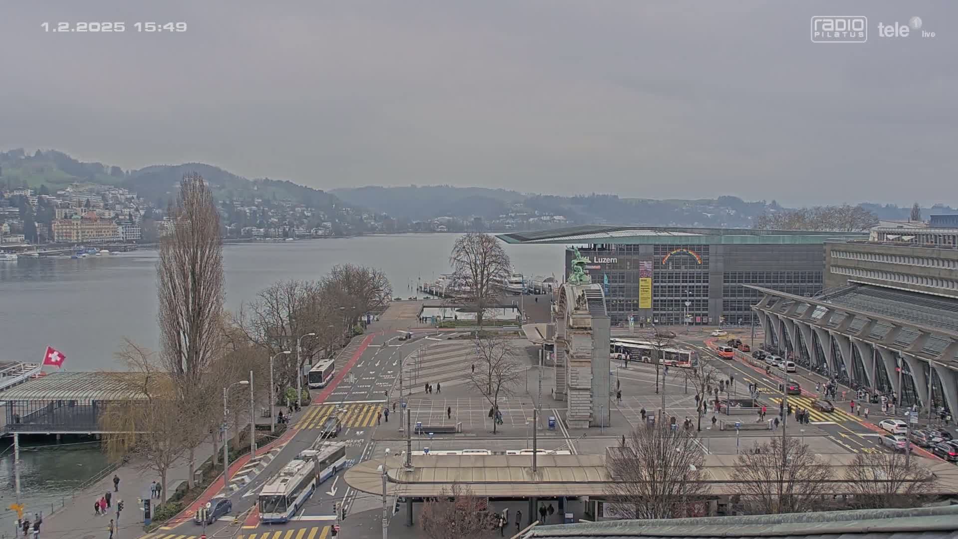 A high-angle, long shot on an overcast day shows a city square near a body of water with buildings, buses, and people;  trees are mostly bare.