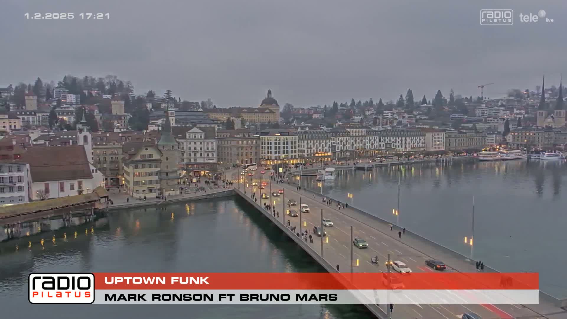 A bridge over a calm river leads to a city of buildings under a gray, overcast sky.