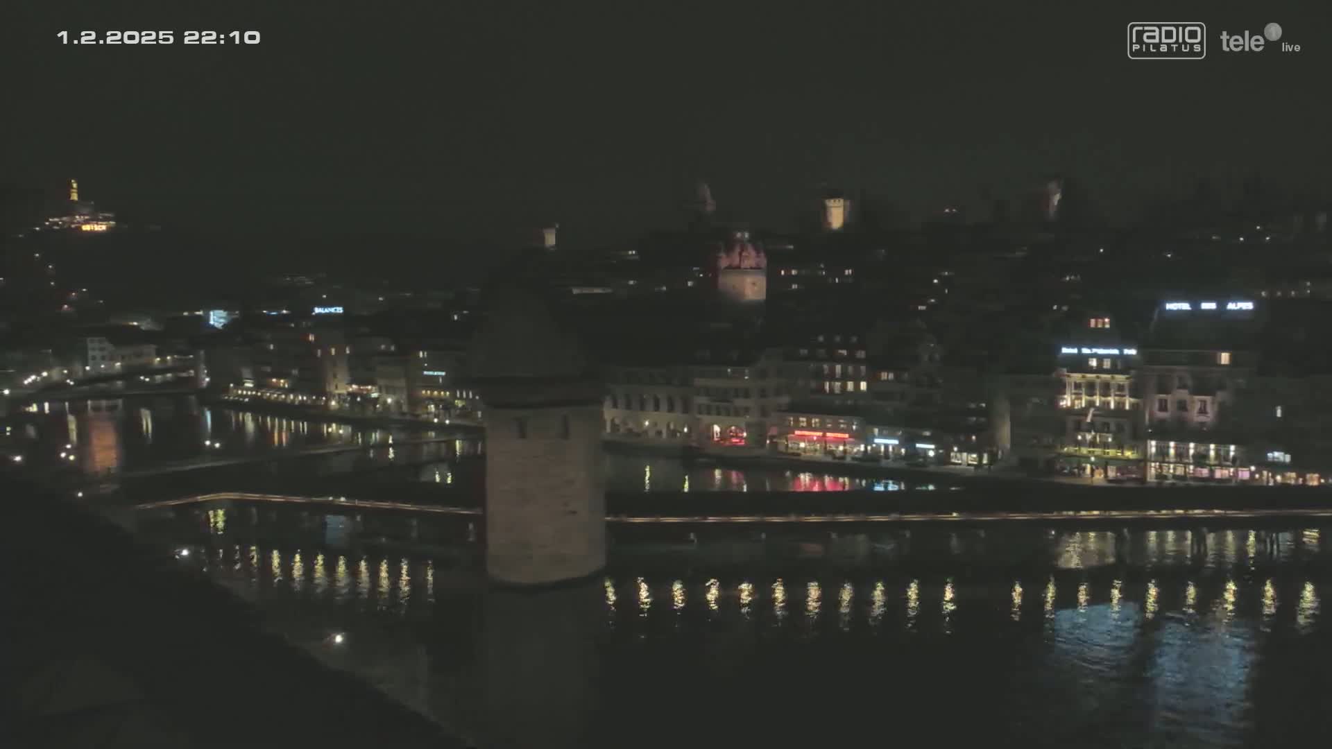 A nighttime view of a city's waterfront, illuminated with lights reflecting on calm water, shows buildings lining the riverbanks.