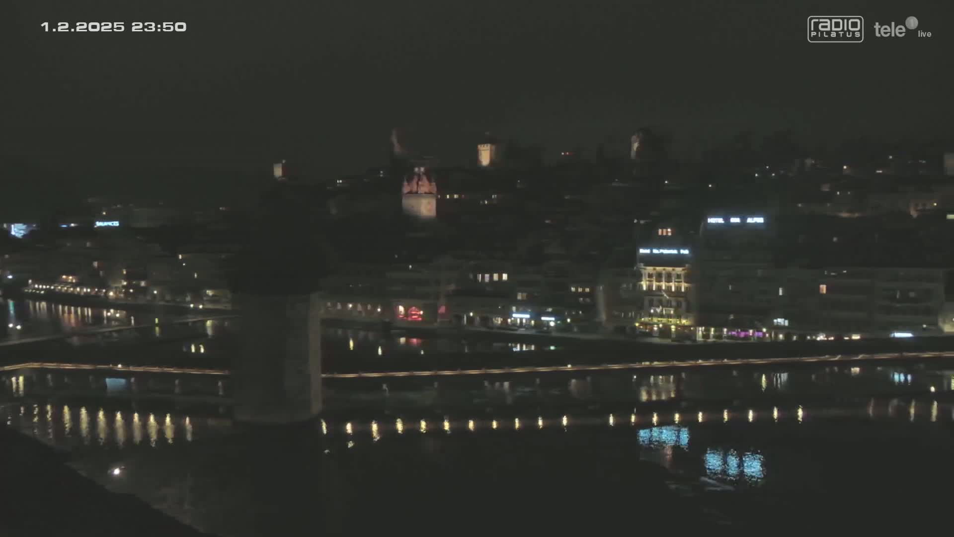 A nighttime, dark view of a city and river, with lights illuminating buildings and reflecting on the water.