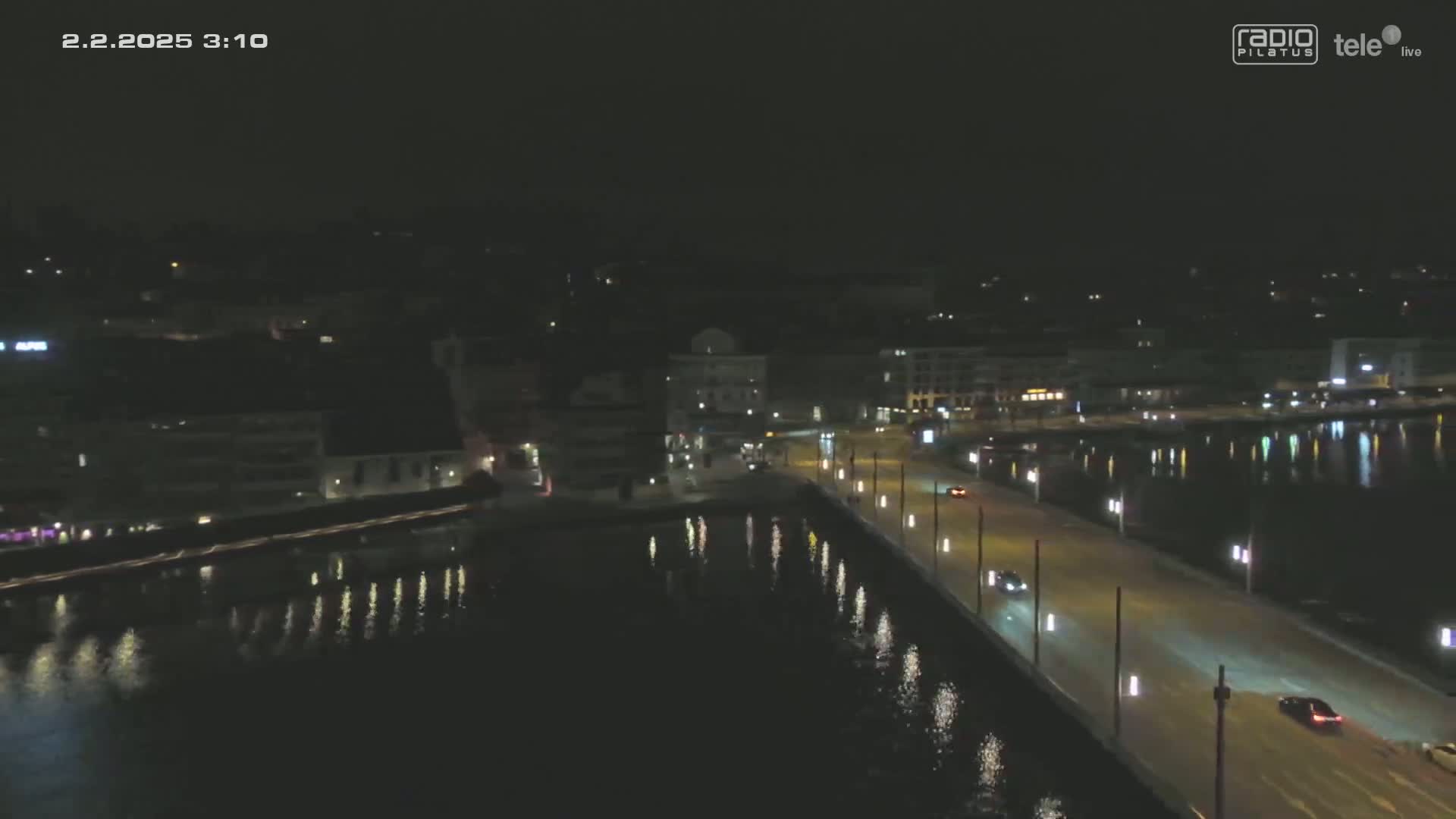 A nighttime, high-angle view shows a city waterfront with a road and few cars, calm water reflecting city lights.