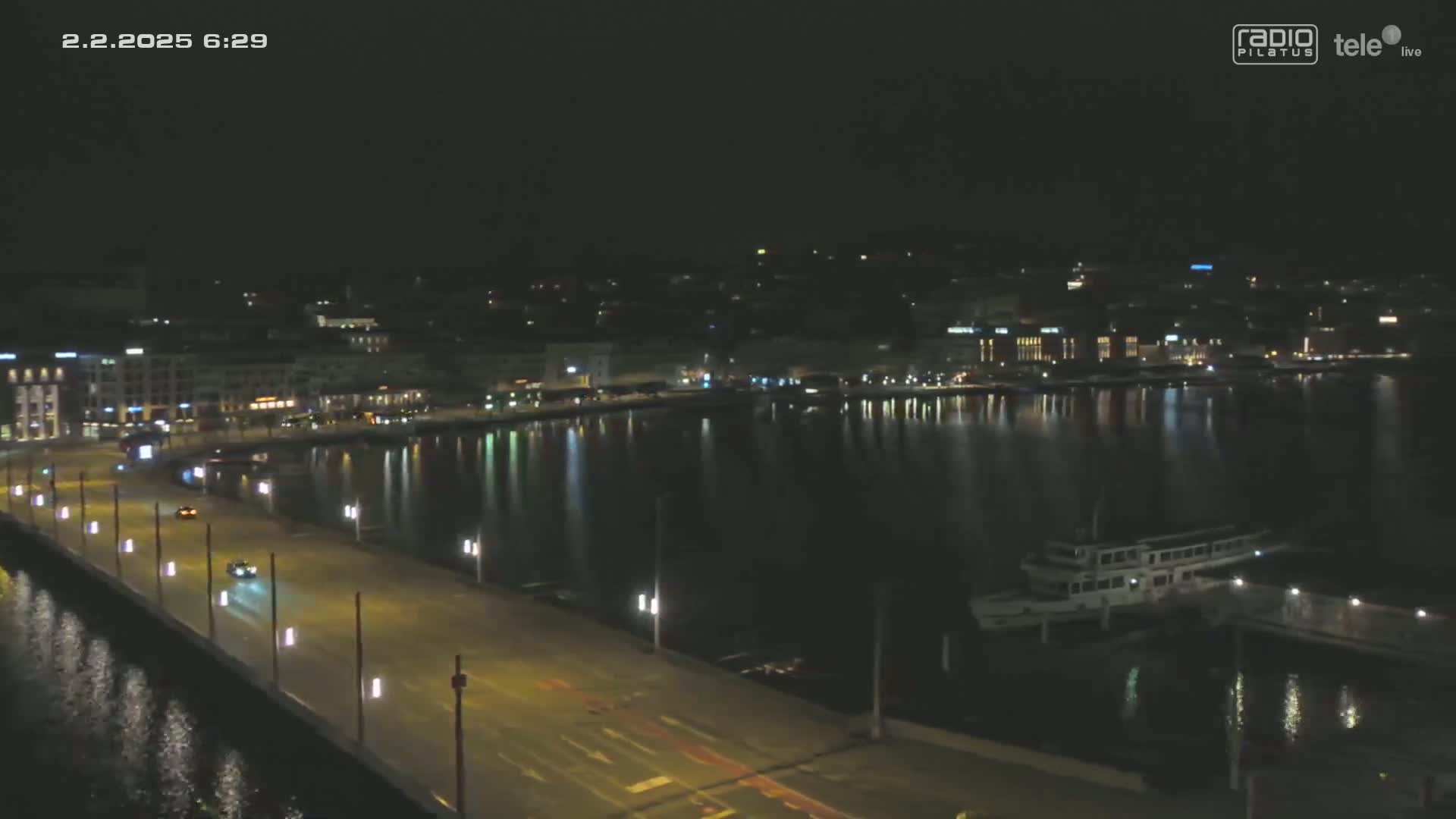 A nighttime view of a city waterfront with a road and a few cars, a body of water reflecting city lights, and a docked boat.
