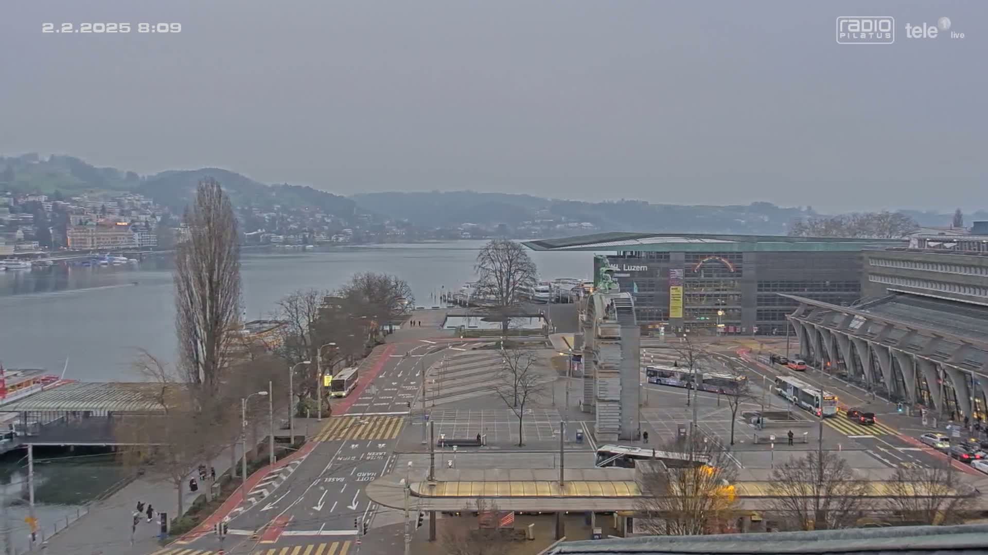 A mostly empty city square overlooks a calm lake and distant hills under an overcast sky.