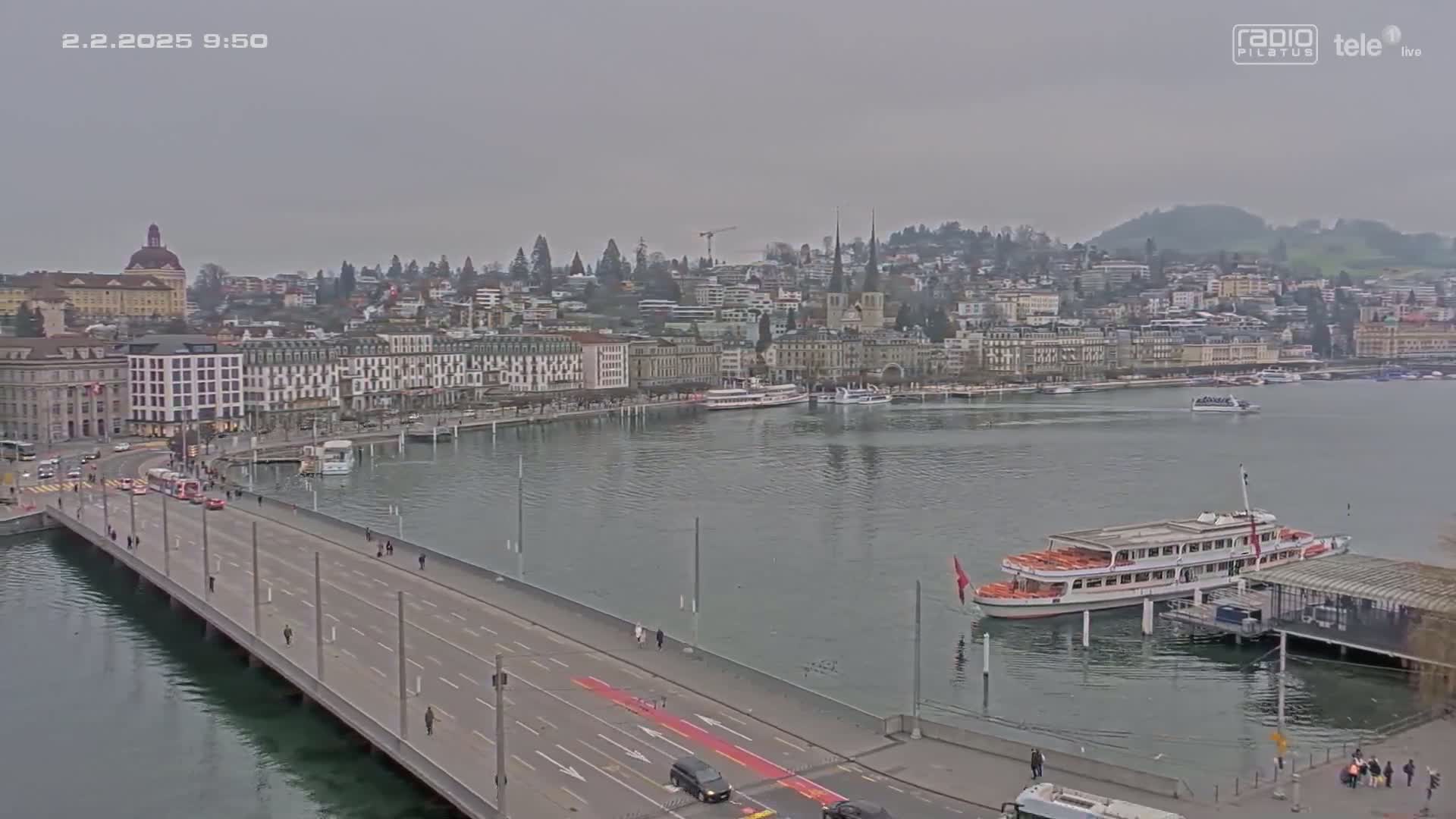 A long bridge stretches over calm water towards a city of buildings on a cloudy day.