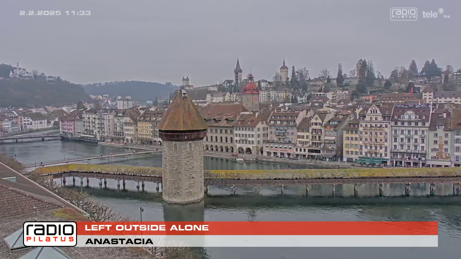 A stone tower stands on a bridge over calm water, in front of a town of closely-packed buildings on a hill, under an overcast sky.