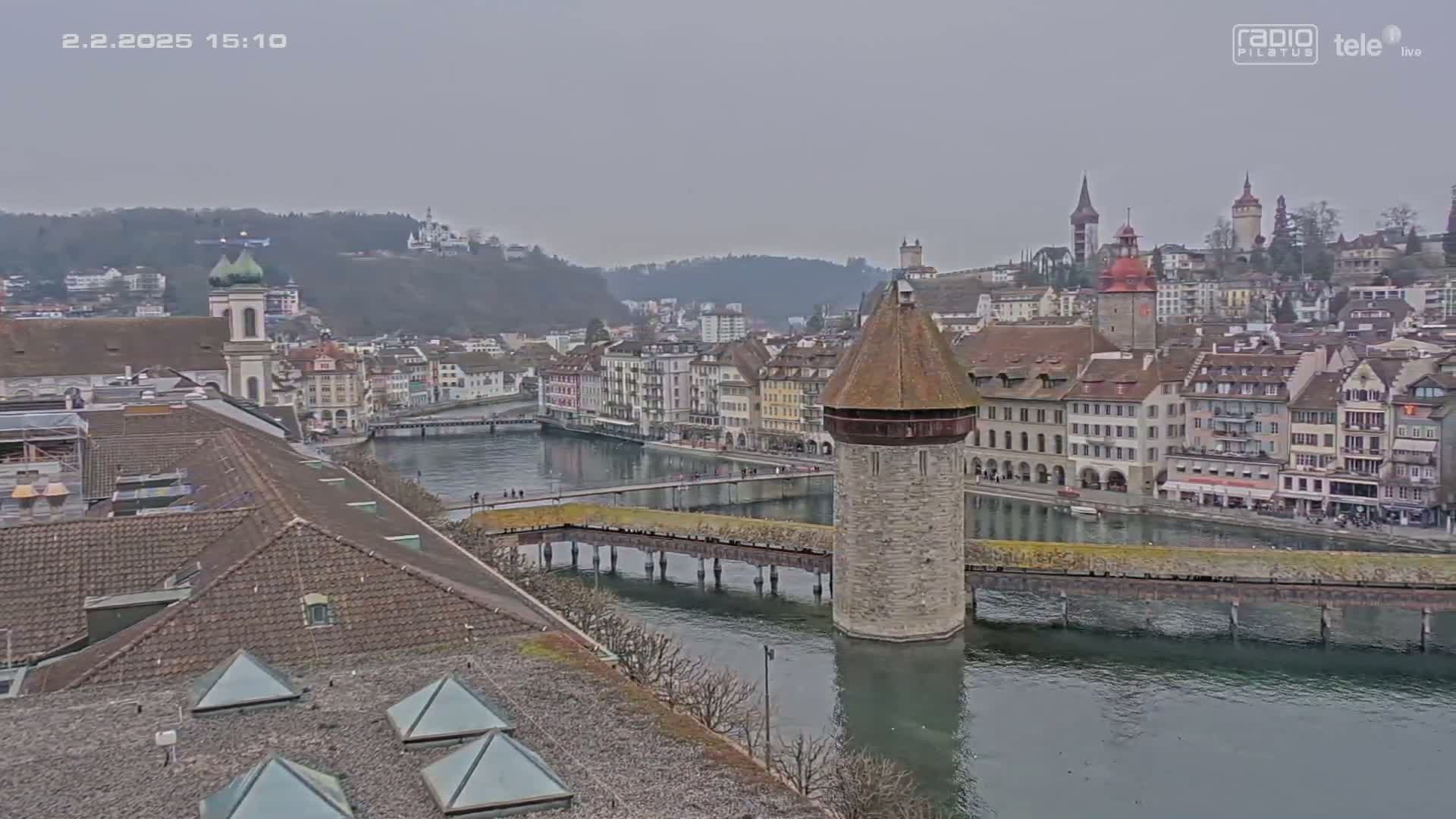 A waterside town is viewed from above on an overcast day, showing a stone tower in the middle of a bridge crossing a river.