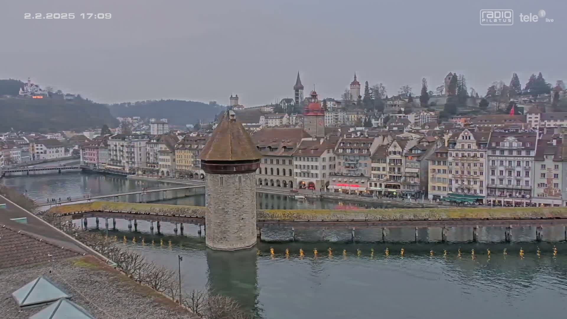 A stone tower stands in a calm river, reflecting in the water, with a quaint town of buildings lining the riverbank under an overcast sky.