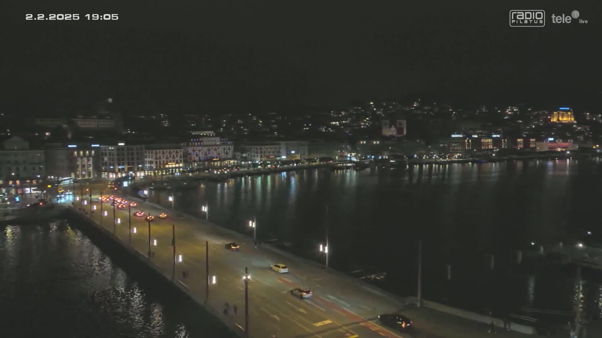 A nighttime aerial view shows a city waterfront with a bridge carrying several cars, under a clear, dark sky.