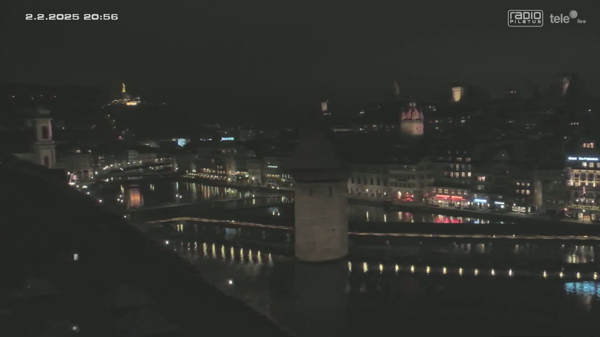 A nighttime view of a city with a river reflecting lights, showcasing buildings along the waterfront and a tower in the middle of the river under a clear, dark sky.