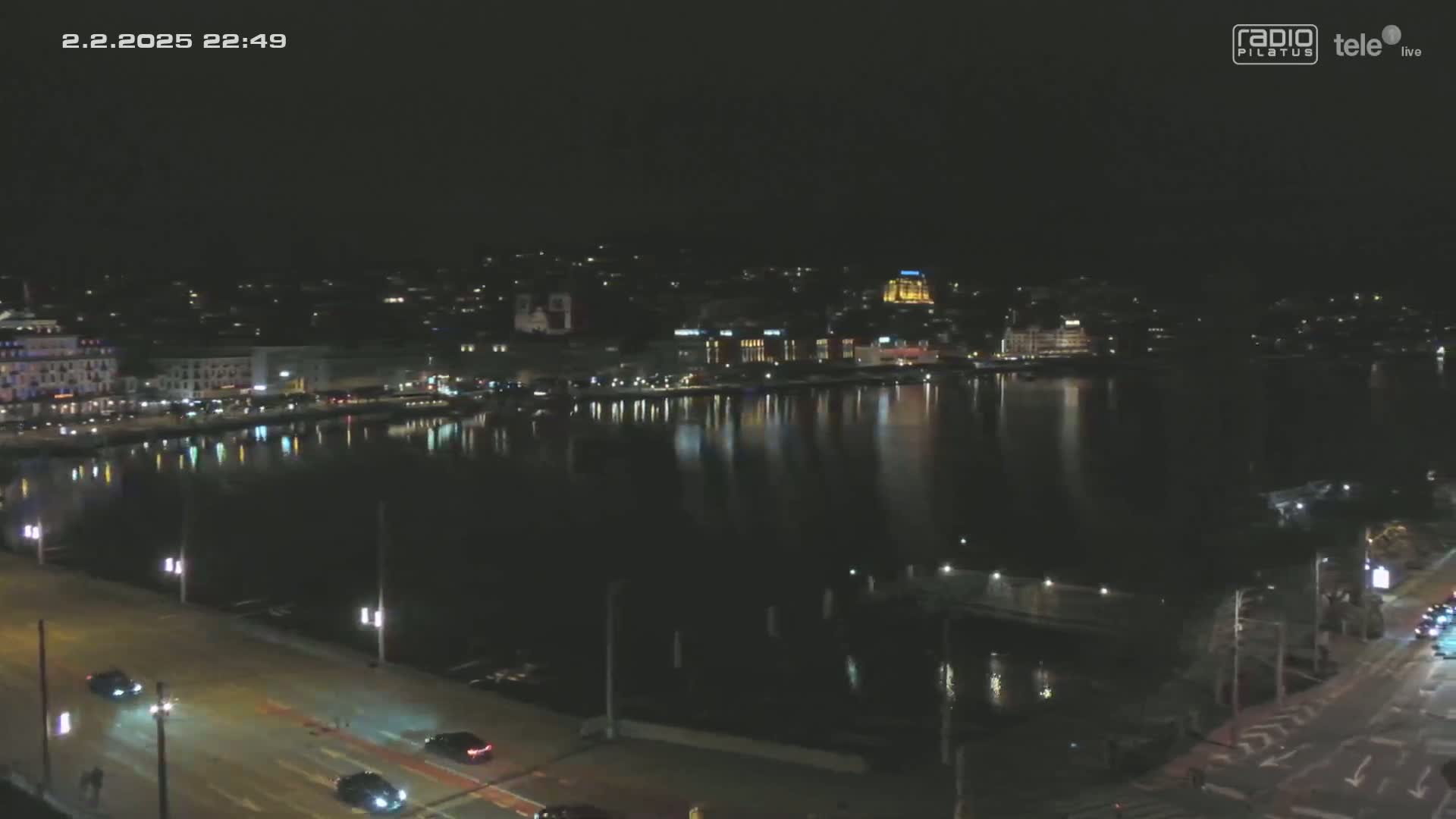 A nighttime view of a city waterfront with calm water reflecting lights from buildings along the shore, and a road with sparse traffic in the foreground.