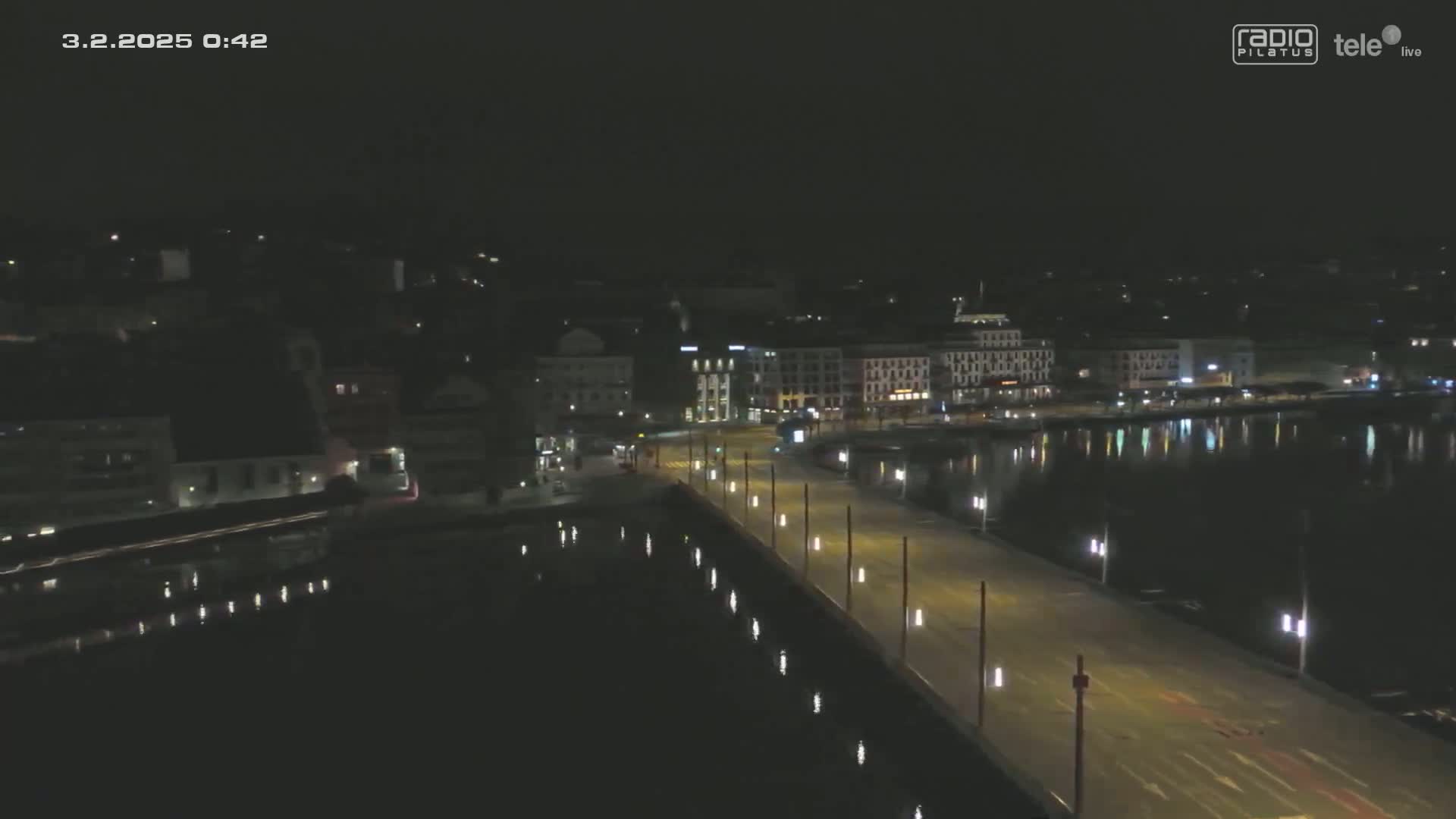 A nighttime, high-angle view shows a waterfront city with a long, illuminated road leading to a body of still water reflecting the city lights.