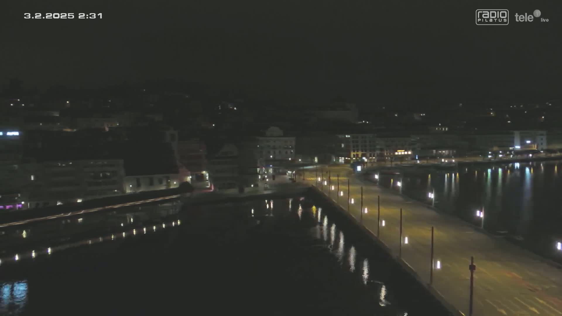 A nighttime, aerial view shows a long, illuminated waterfront walkway extending into a calm, dark body of water, alongside a city with dimly lit buildings.