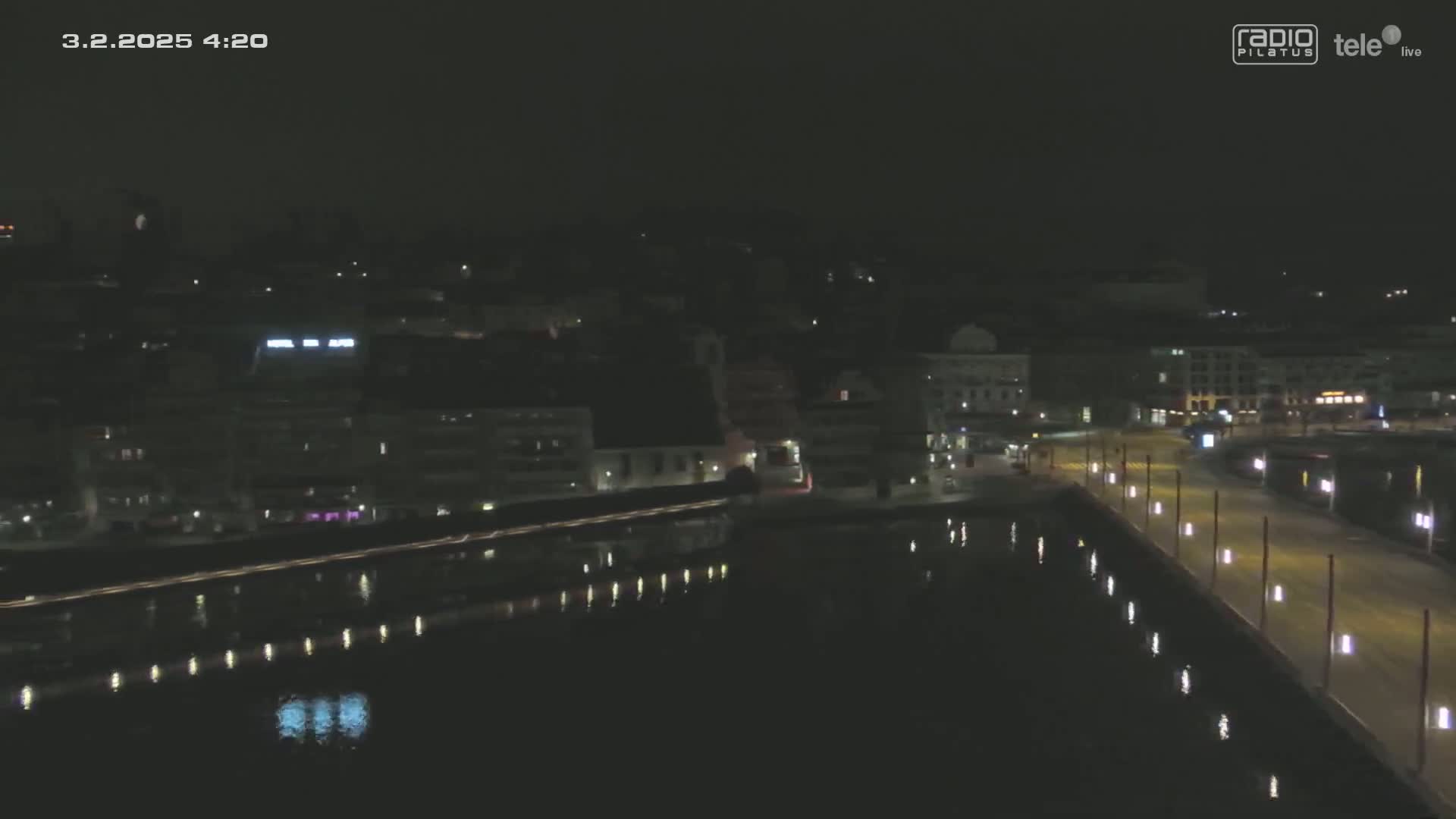A nighttime aerial view shows a city waterfront with calm water reflecting lights from buildings and streetlamps along a roadway.