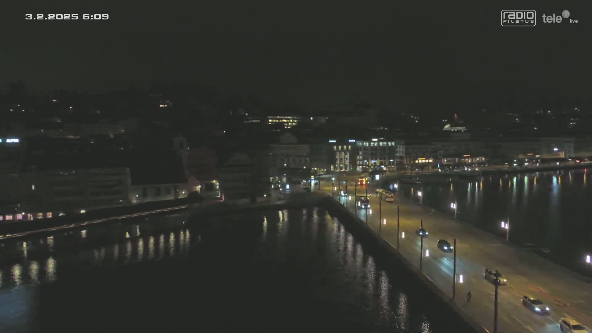 A nighttime, high-angle view shows a bridge with cars driving across it, alongside a dark body of water reflecting the city lights.