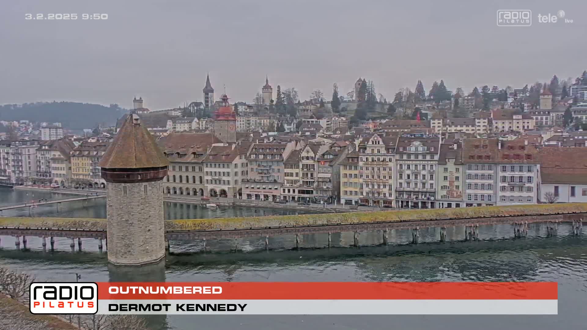 A view of a city with numerous buildings along a waterfront, under an overcast sky, features a stone tower situated on a bridge extending into the water.