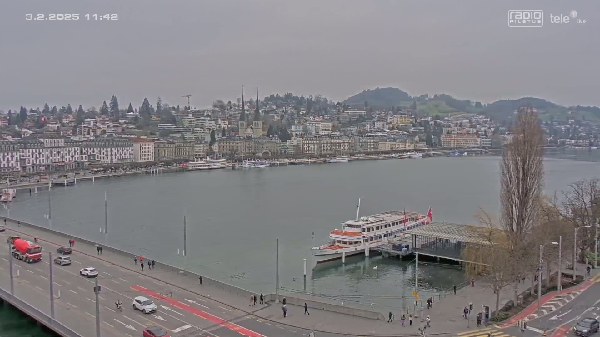 A city waterfront with many buildings along the shore, a large passenger boat docked, and a road with cars driving on a cloudy day.