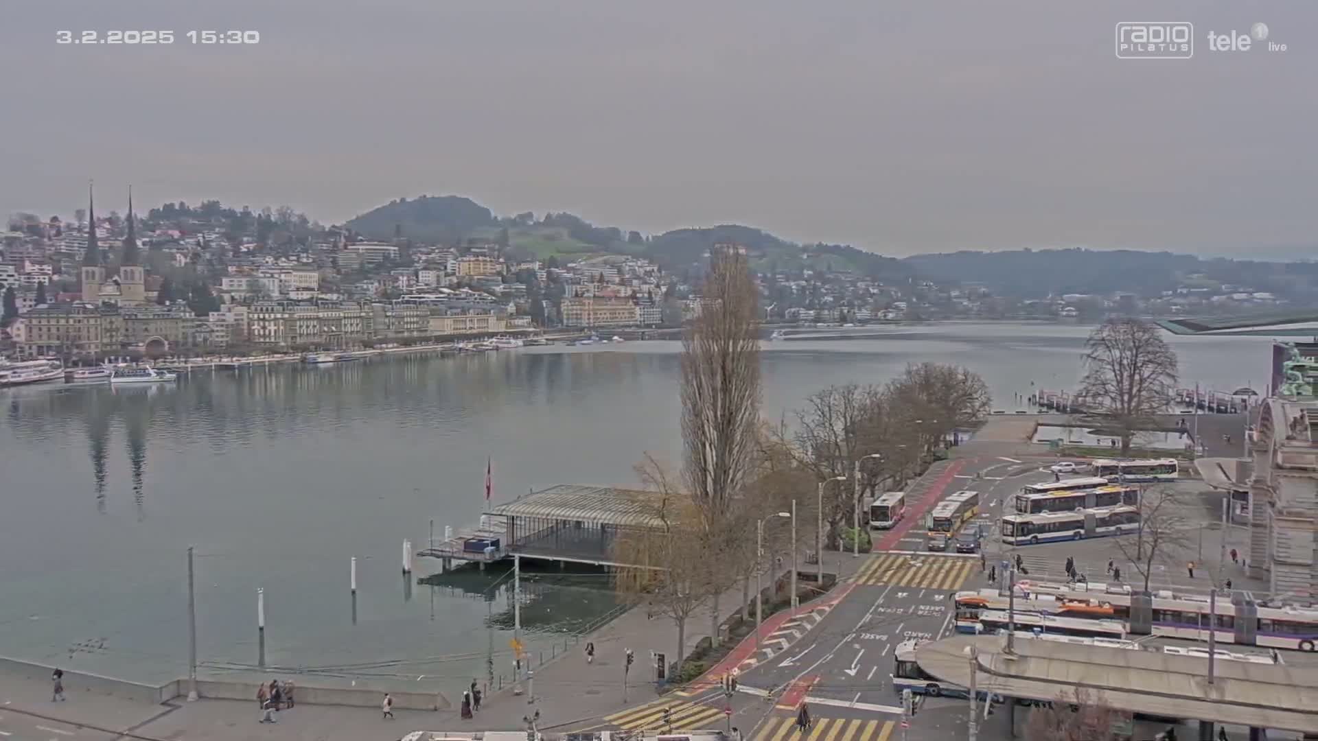 A waterfront city scene on an overcast day shows a calm lake, a city skyline with buildings on a hill, and buses at a terminal near the water's edge.
