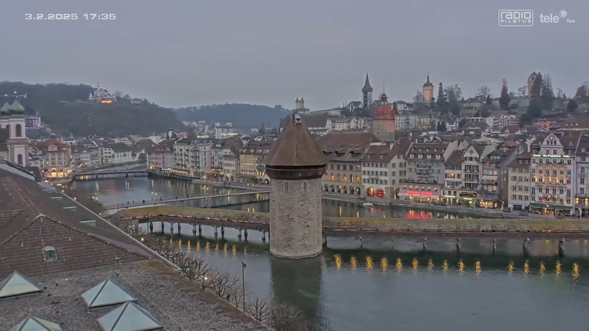 A twilight view of a river flowing through a city with a stone tower in the middle of a bridge, surrounded by buildings with lights on.