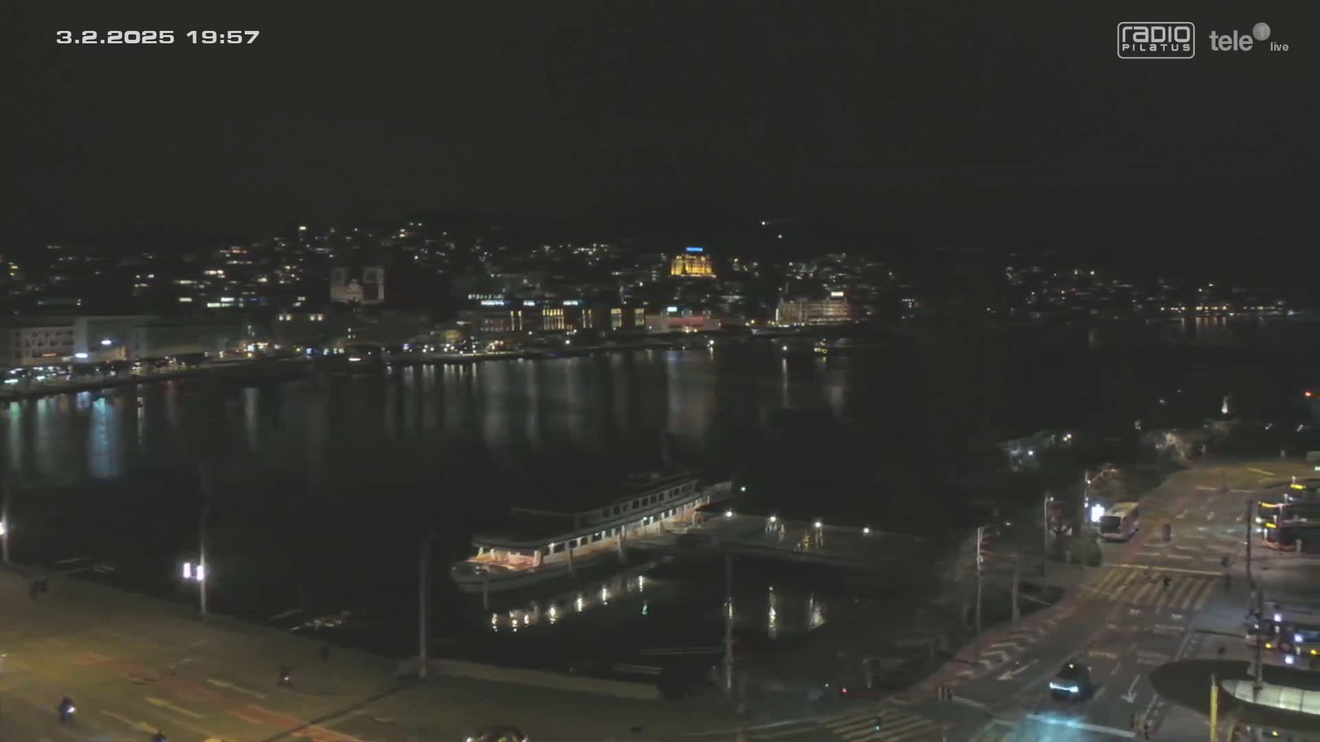 A nighttime view of a city waterfront with a body of calm water reflecting city lights, a docked boat, and illuminated streets below.