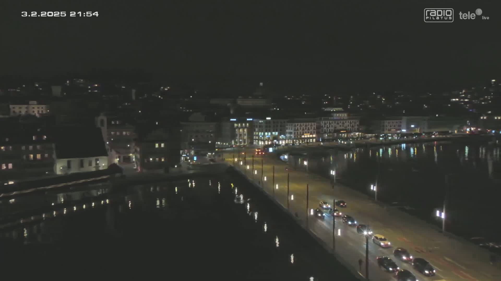 A nighttime aerial view shows a city waterfront with a road lined with cars and lights reflecting on dark water.