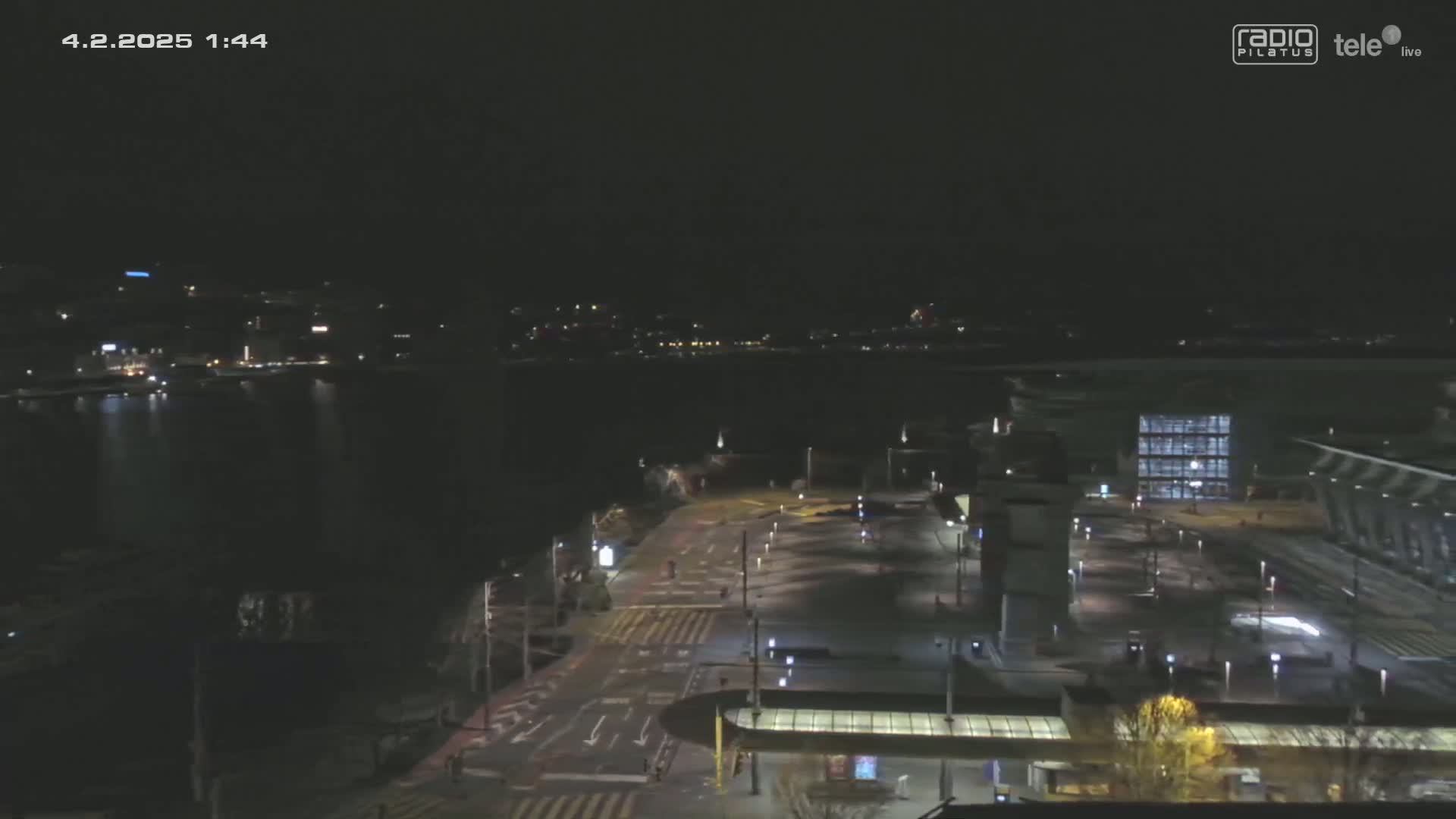 A nighttime, high-angle view shows a mostly empty city square and roadway next to a body of water with city lights visible in the distance.