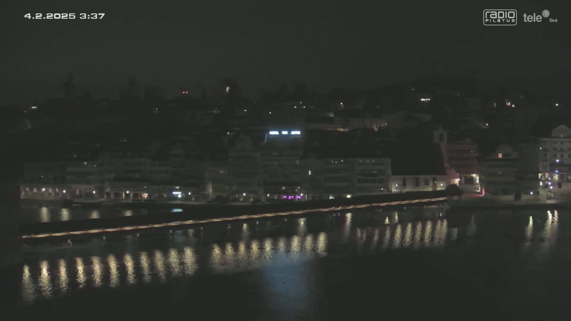 A nighttime view of a town situated on a body of water, featuring a bridge illuminated with lights, and the lights of buildings reflecting on the calm water.