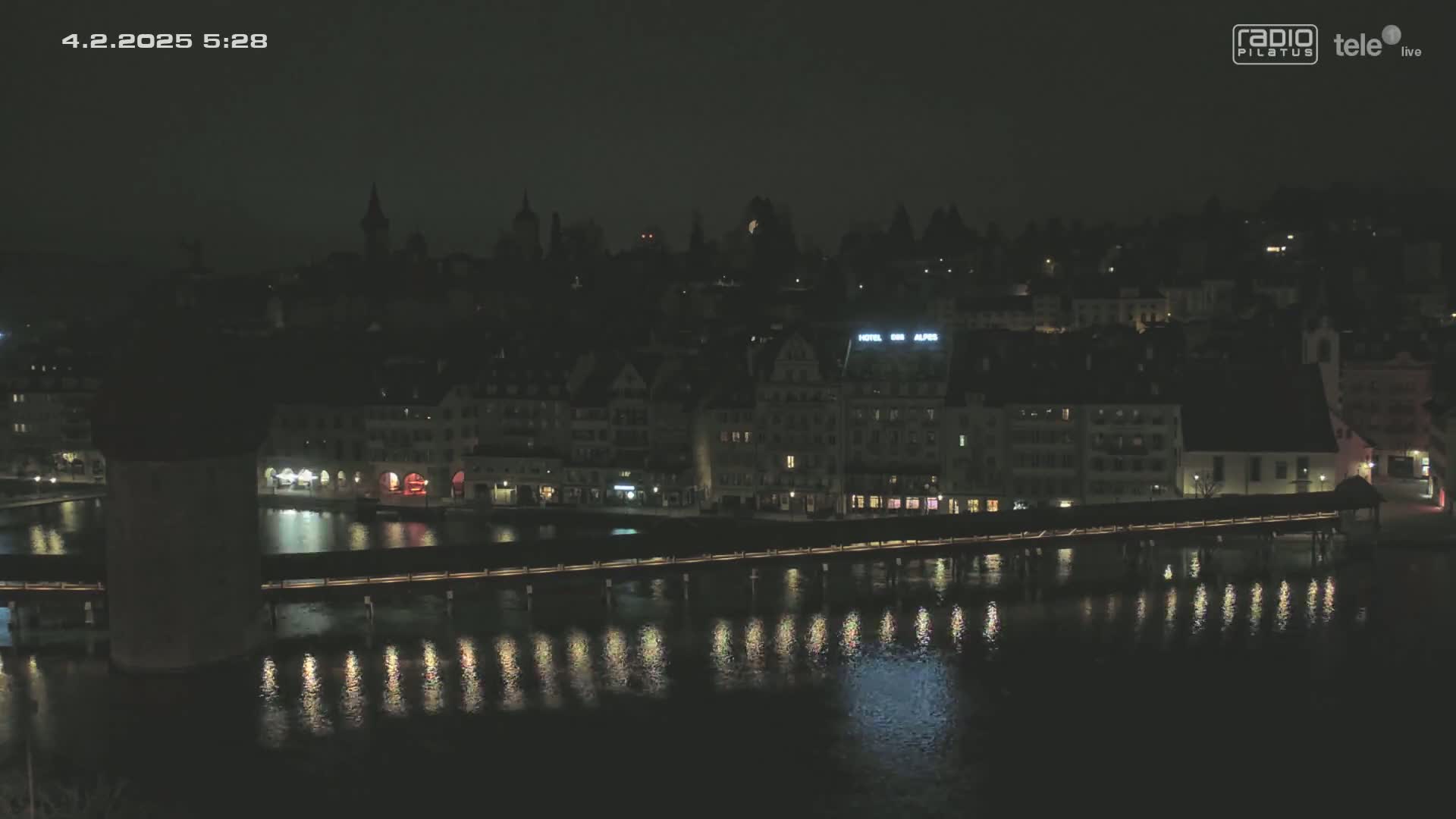 A nighttime view of a town with a bridge spanning a body of water, with the lights of buildings and their reflections shimmering on the water's surface.