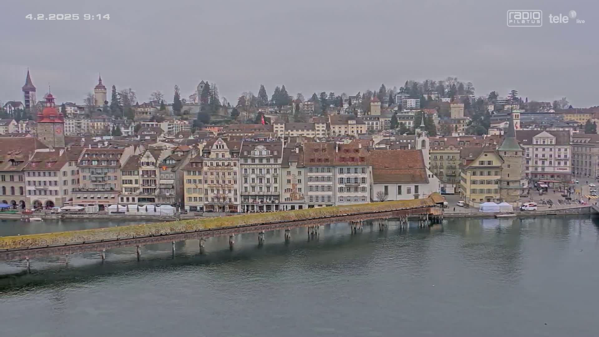 A long, covered bridge spans a calm body of water in front of a European city on an overcast day.