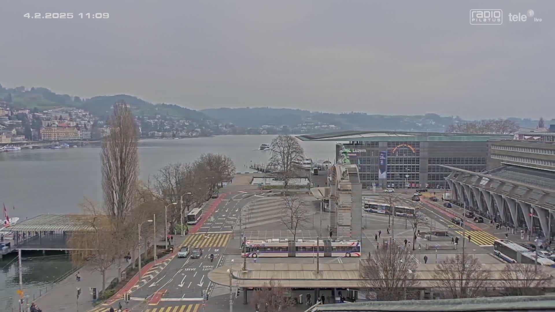 A cloudy day overlooking a city's waterfront with a body of water, buildings, vehicles, and sparsely leaved trees.