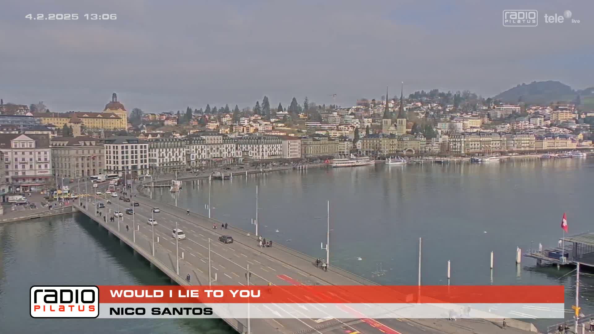 A bridge over a calm lake leads to a city with many buildings under a cloudy sky.