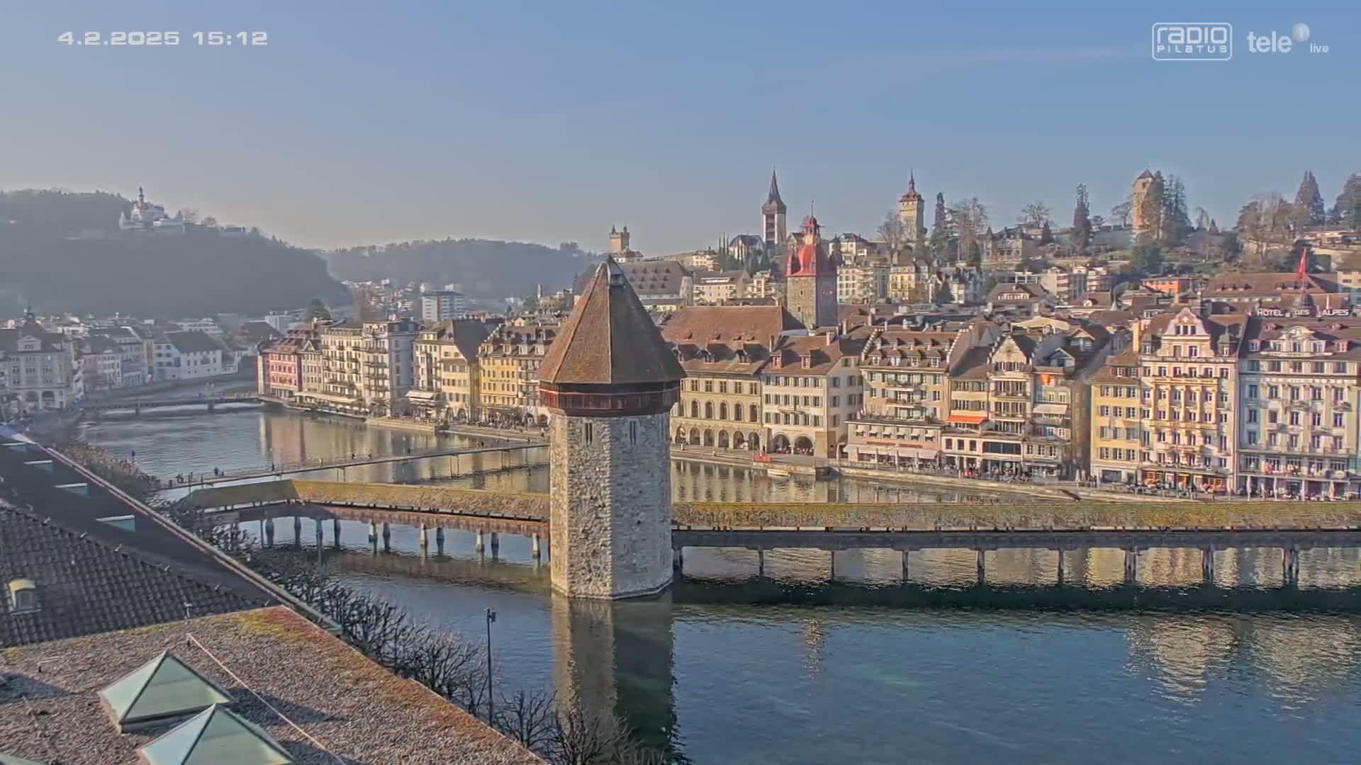A stone water tower stands in a calm river, connected to a long wooden bridge spanning the water to a town of colorful buildings under a clear, sunny sky.