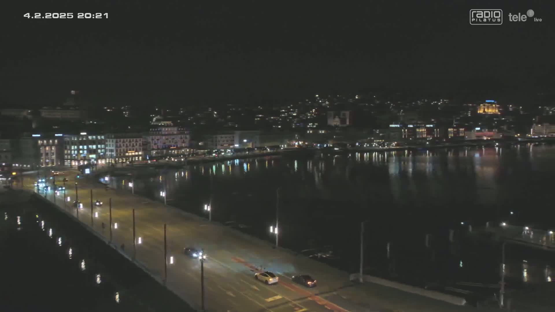 A nighttime, high-angle view shows a city waterfront with a bridge and calm water reflecting city lights.