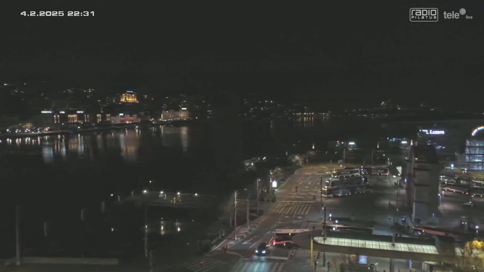A nighttime view of a city across a body of water, with city lights reflecting on the water's surface and traffic visible on roads near a waterfront bus terminal.
