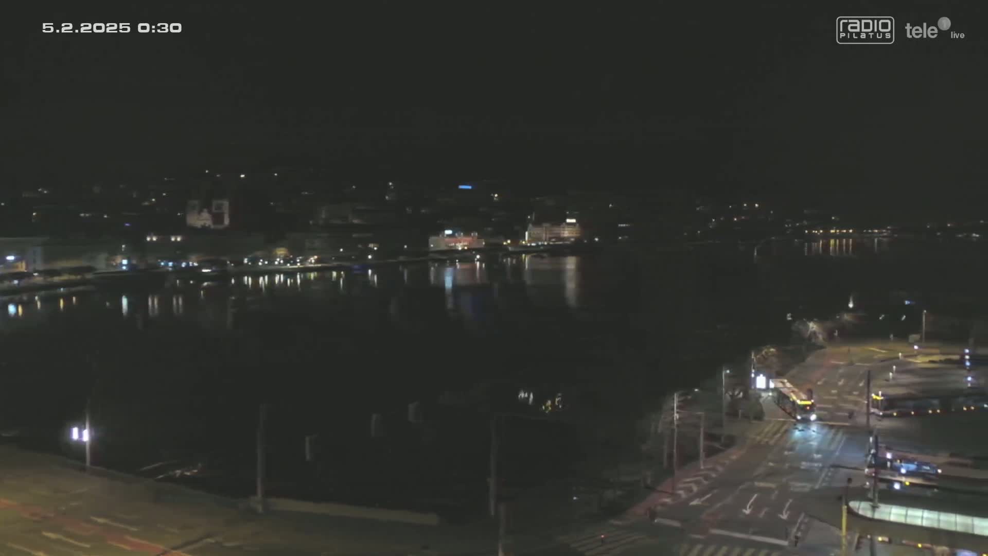 A nighttime, city waterfront scene shows a body of water reflecting city lights, with streets and vehicles visible in the foreground.