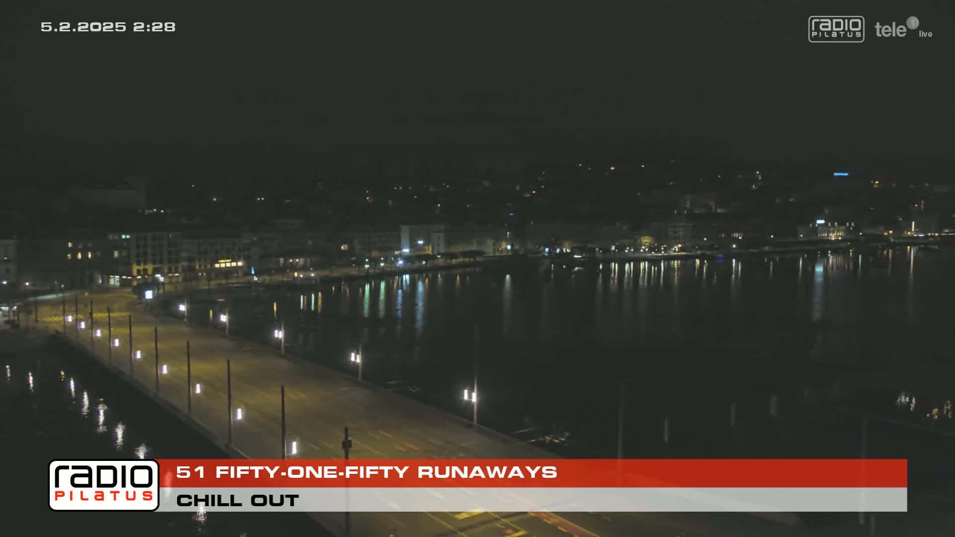 A nighttime, long shot of a city waterfront with a bridge, calm water reflecting the city lights.