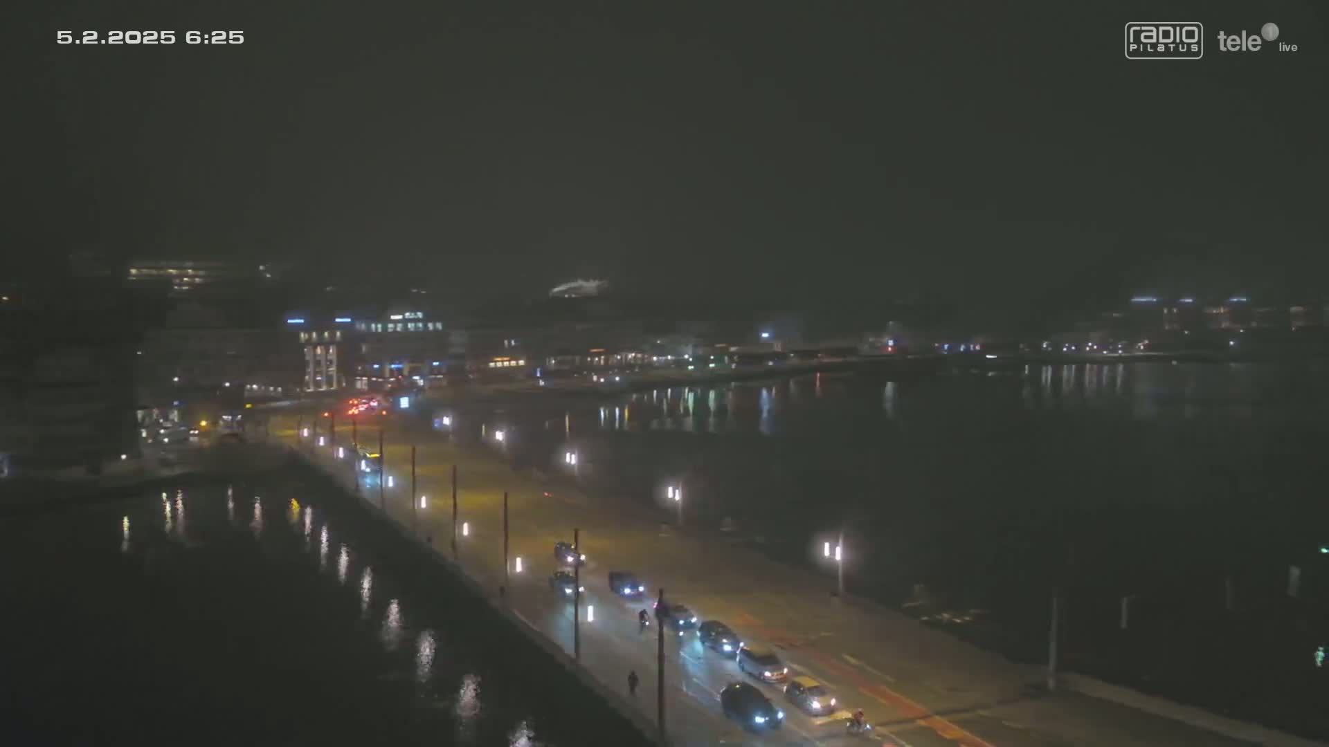 A nighttime view of a city waterfront shows a road with cars driving along it, next to a body of dark water reflecting city lights under a dark sky.